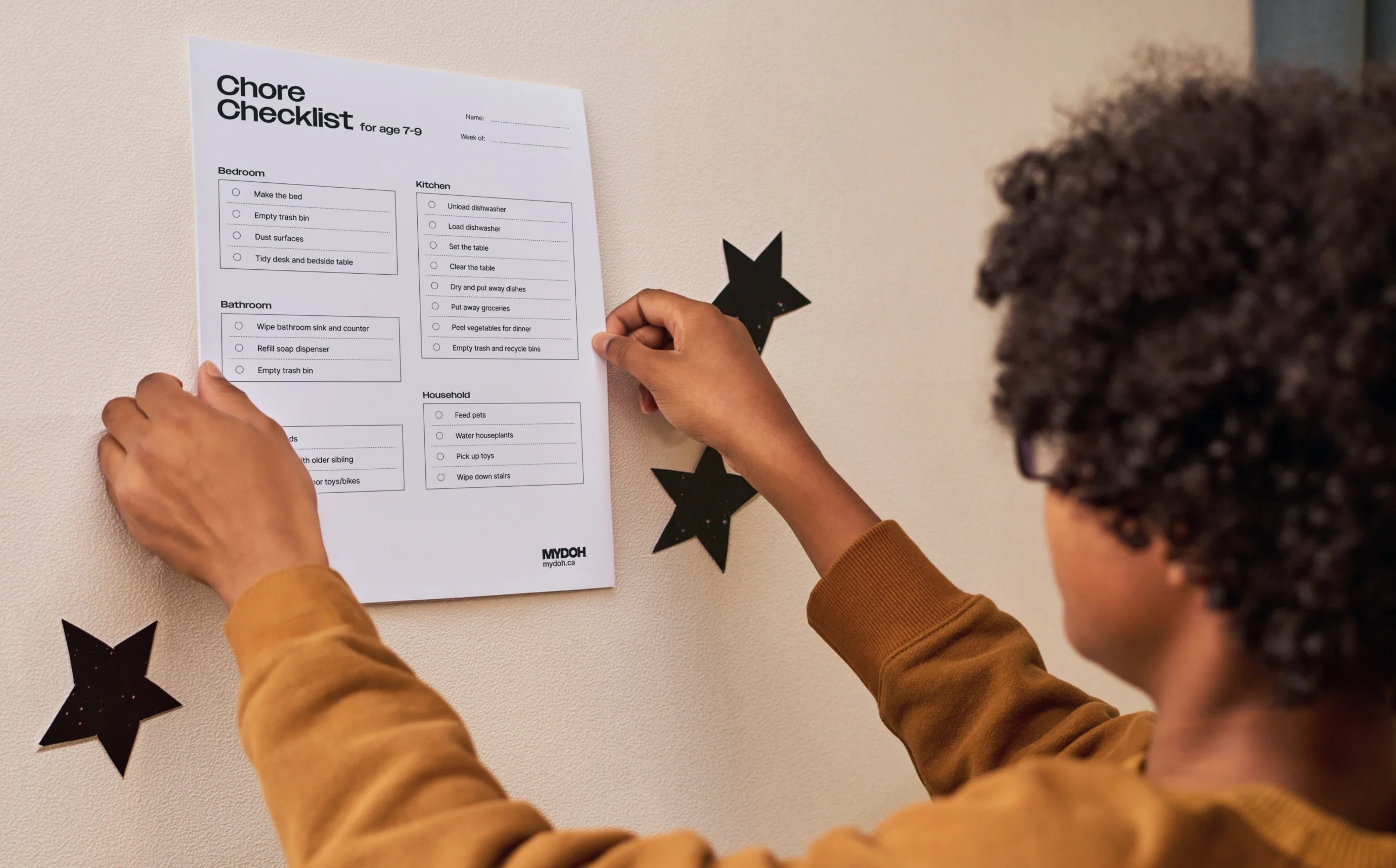 A child in a yellow sweatshirt mounts a Chore Chart checklist for ages 7 to 9 on a cream wall decorated with black star decals.