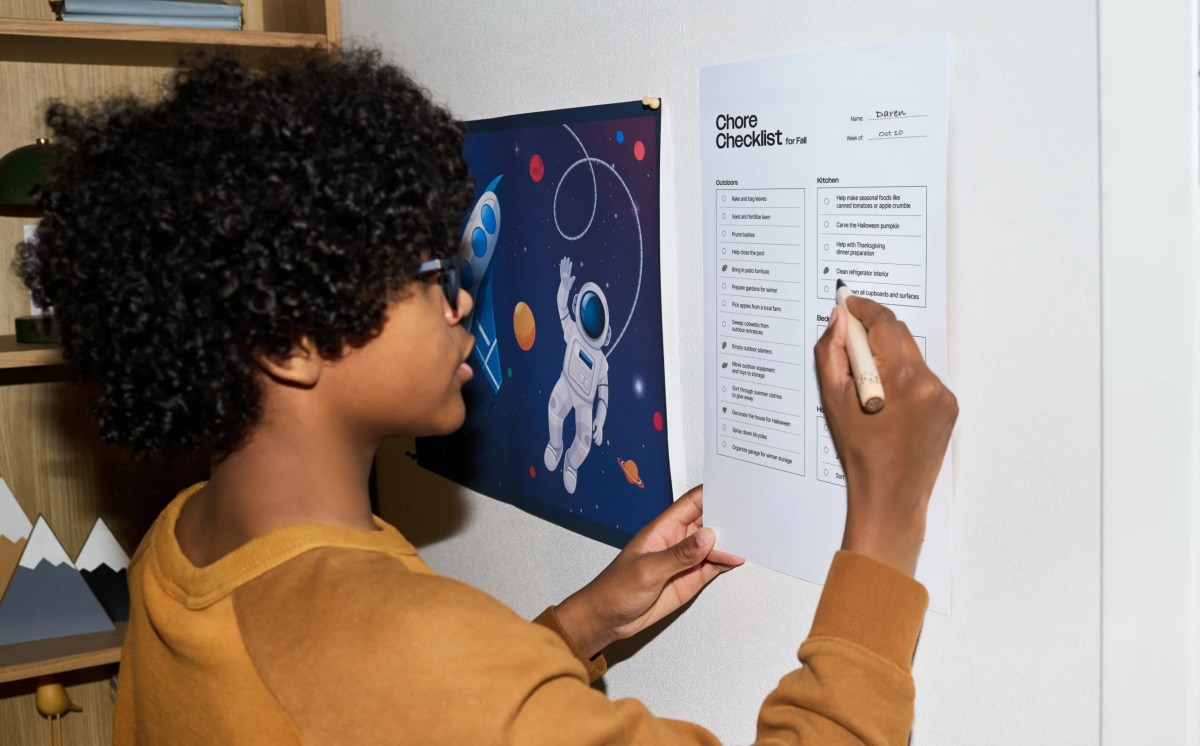A child in a yellow sweatshirt checks off chores on a Fall Chore Chart checklist pinned to a bedroom wall beside a space poster.