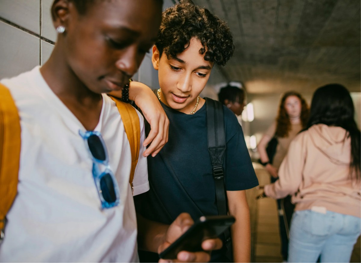 Two students lean together looking at a smartphone in a tiled school hallway, backpacks on, while others walk past in the background.