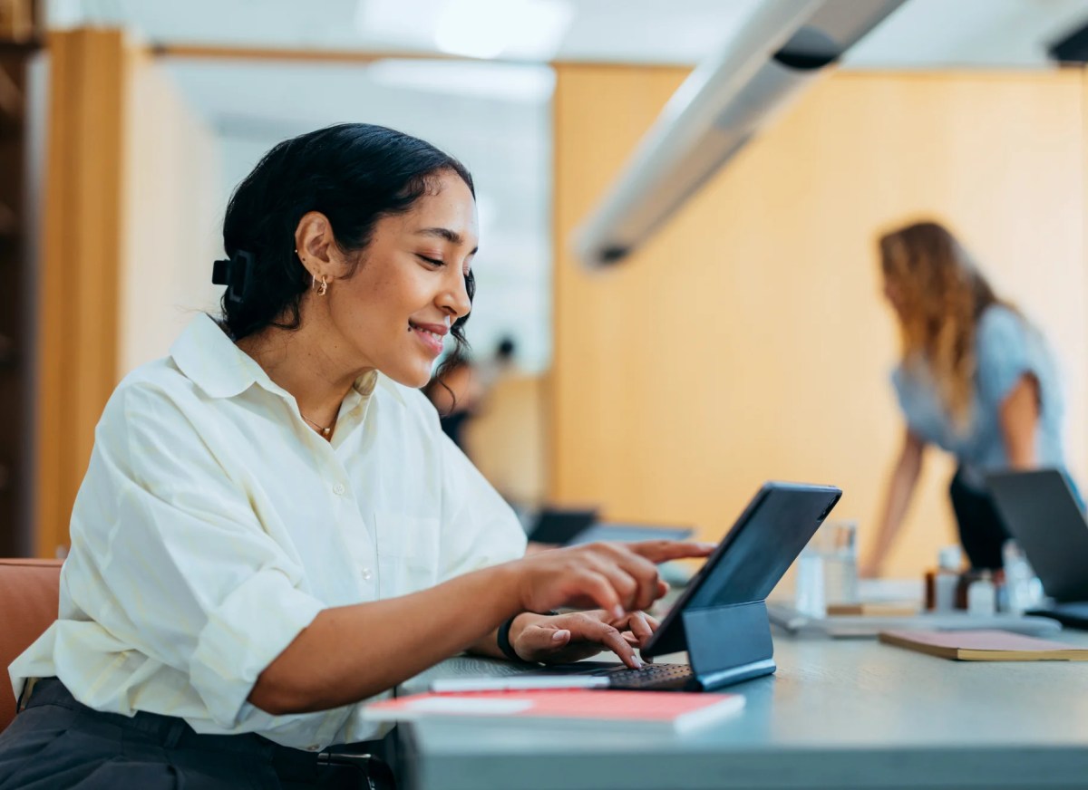 A person working on a tablet with keyboard in a bright modern office, smiling while touching the screen during a productive career moment.